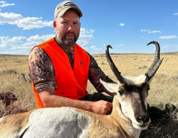 A hunter sitting next to a pronghorn antelope in Wyoming's dry terrain, wearing an orange safety vest and camouflage, representing a memorable hunt guided by SNS Outfitter & Guides.